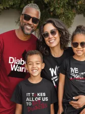 A family of four stands in front of a leafy green wall. The father wears a dark red t-shirt that says "Diabetes Warrior" in white letters. The mother wears a black t-shirt that says "We Fight" in white letters. The young son wears a black t-shirt with the words "It Takes All of Us to Stop Diabetes" written on it. The young daughter wears a black t-shirt with the words "We Fight" in white letters. All are smiling and wearing sunglasses.